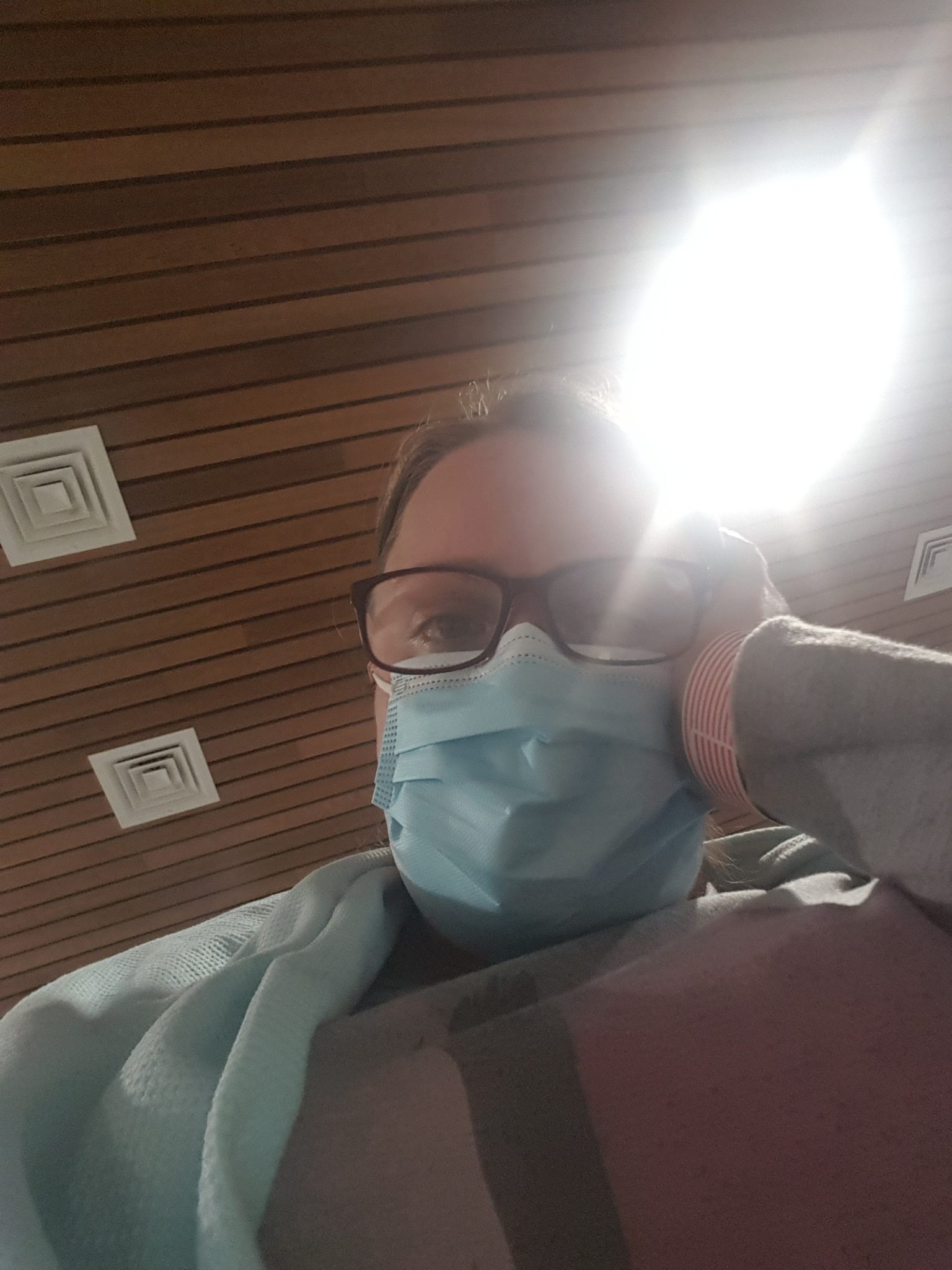 [Image Description: Ellie takes a selfie from below. She has a grey top on, a blue blanket around her shoulder and a blue mask on her face. She has glasses on and her head resting on her hand. In the background is a wood-plank ceiling with a light shining brightly.]