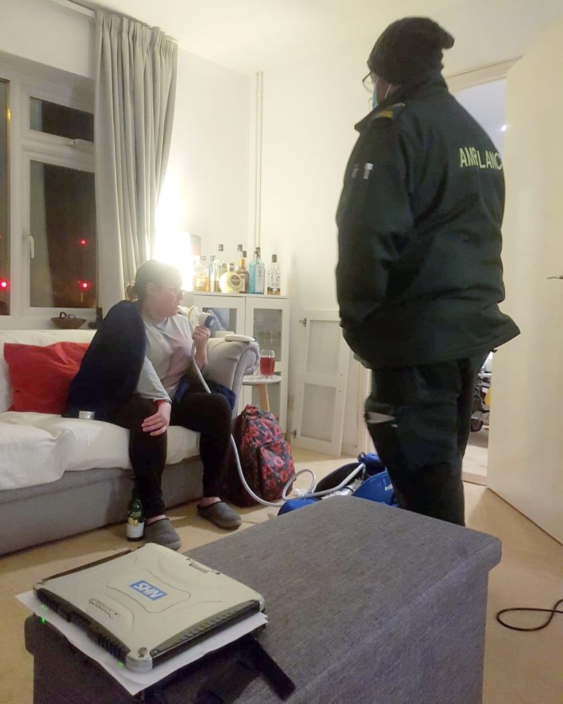 [Image description: Ellie sits on the sofa in a living room with windows in the background being administered entonox via a tube to her mouth from a bag on the floor. In the middle of the picture a paramedic stands with his back to the camera and in the foreground a grey coffee table has a light grey laptop on it with an NHS sticker on it].